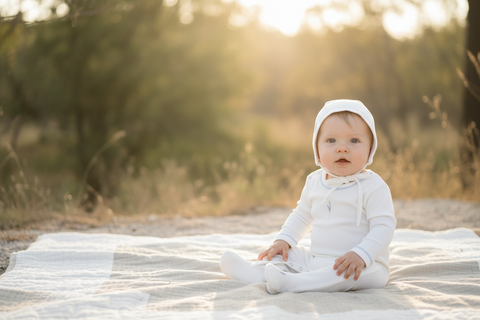 Baby in a white outfit sitting on a blanket outdoors with a blurred natural background